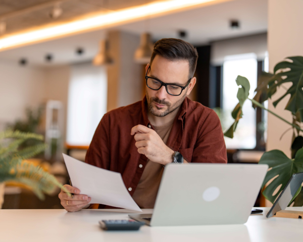 Mann mit Brille prüft Unterlagen am Laptop in modernem Büro.