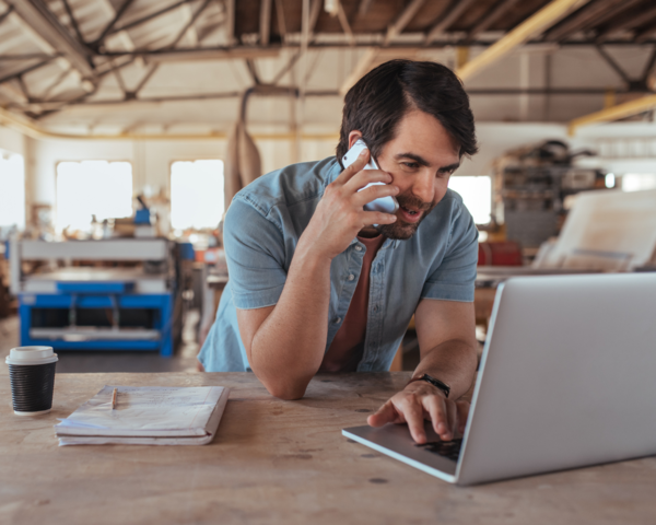 Business-Telefonie im Arbeitsalltag Mann telefoniert im Betrieb und arbeitet parallel am Laptop.