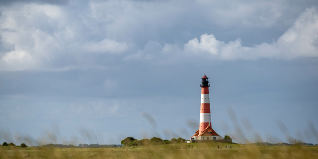 Ein rot-weiß gestreifter Leuchtturm steht in einer weiten Graslandschaft unter einem wolkigen Himmel.