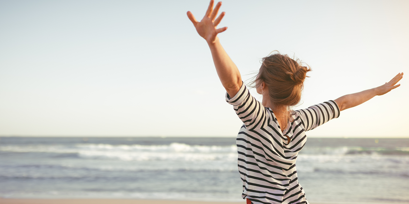 Eine Frau steht am Strand mit offenen Armen Richtung Horizont – Symbolbild für Freiheit, Leichtigkeit und Flexibilität. Ideal für die Darstellung der Vorteilswelt rund um unsere Glasfaser-Tarife.