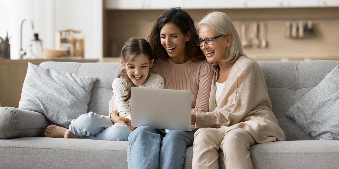 Eine Großmutter, Mutter und Tochter sitzen gemeinsam auf einem Sofa und lachen, während sie auf einen Laptop schauen. Das Bild symbolisiert digitale Teilhabe, moderne Kommunikation und wie Glasfaser Menschen jeden Alters miteinander verbindet.