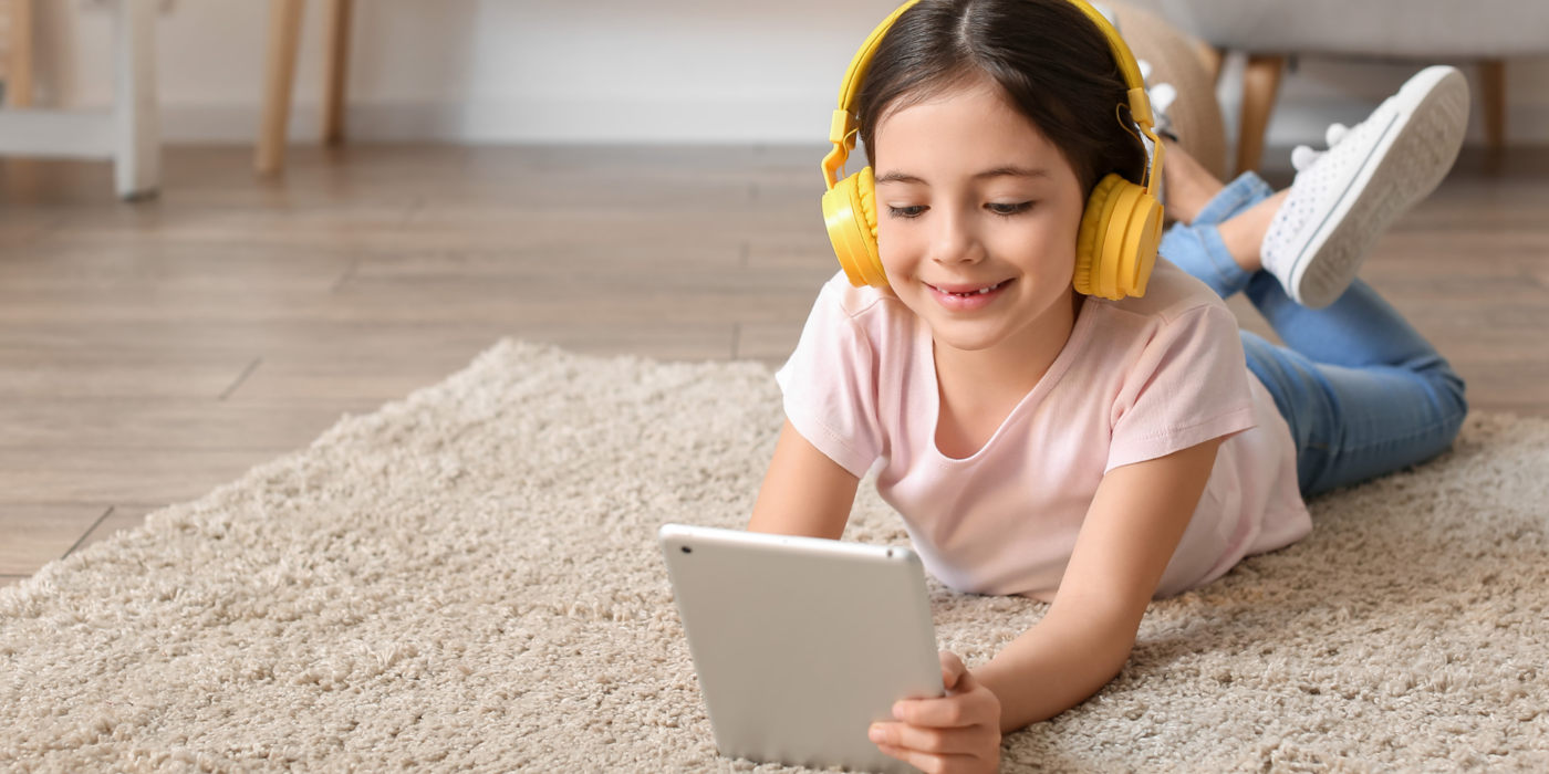 Ein Kind liegt entspannt auf dem Teppichboden und schaut mit Kopfhörern ein Video auf dem Tablet – ein Sinnbild für sorgenfreies und geschütztes Surfen mit dem Sicherheitspaket im Internet.