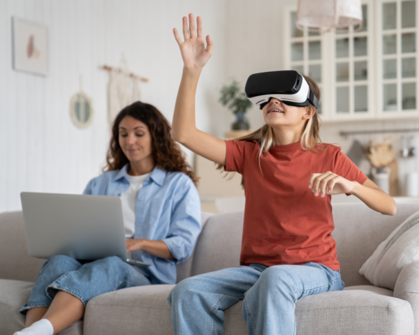 Frau mit Laptop und Kind mit VR Brille sitzen auf dem Sofa