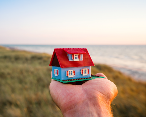 Hand hält Miniaturhaus vor Küste bei Sonnenuntergang