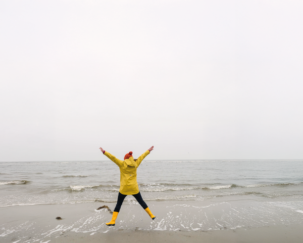 Frau in gelber Regenjacke springt am Strand vor dem Meer