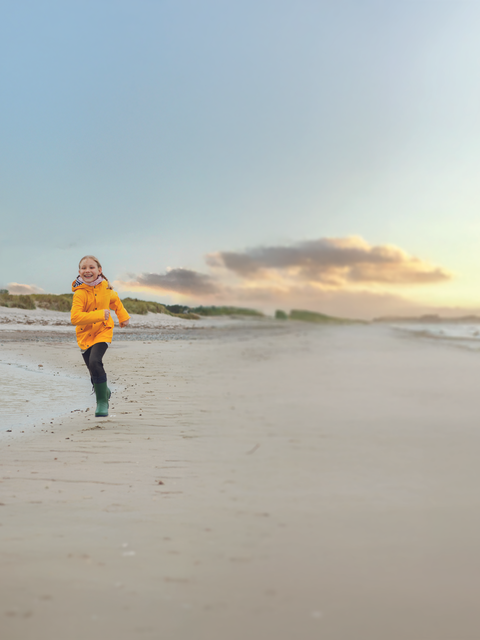 Ein Kind läuft fröhlich am Strand entlang, während im Hintergrund ein Leuchtturm steht.