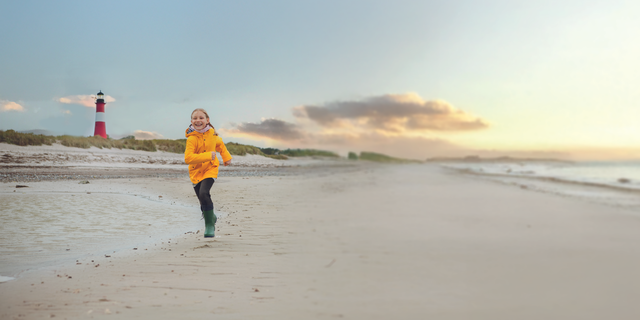 Ein Kind läuft fröhlich am Strand entlang, während im Hintergrund ein Leuchtturm steht.