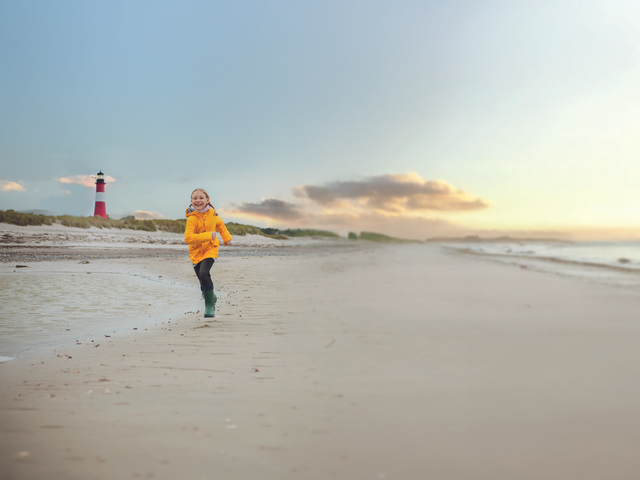 Kind am Strand Ein Kind läuft fröhlich am Strand entlang, während im Hintergrund ein Leuchtturm steht.