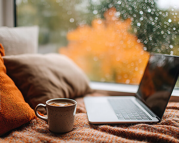 Ein Laptop und eine Tasse mit Getränk liegen auf einer Decke am Fenster, während draußen buntes Herbstlaub und Regentropfen zu sehen sind.